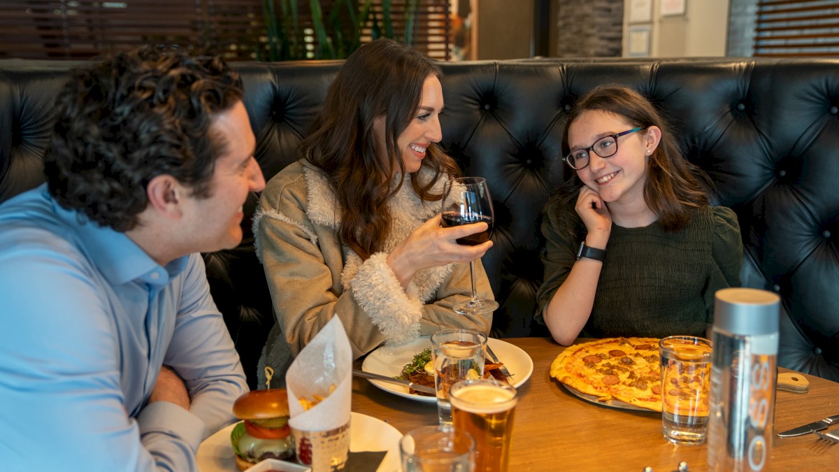 A family of three is dining at a restaurant, enjoying a meal together with burgers, pizza, and drinks, engaging in conversation and smiling.