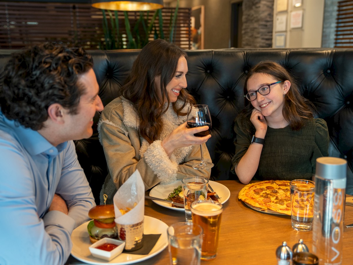 A family of three is dining at a restaurant, enjoying a meal together with burgers, pizza, and drinks, engaging in conversation and smiling.