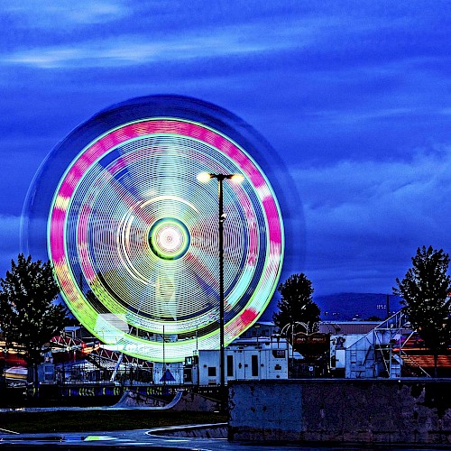 A brightly lit amusement park at night with a spinning Ferris wheel, other rides, and trees under a dark blue sky ending the sentence.