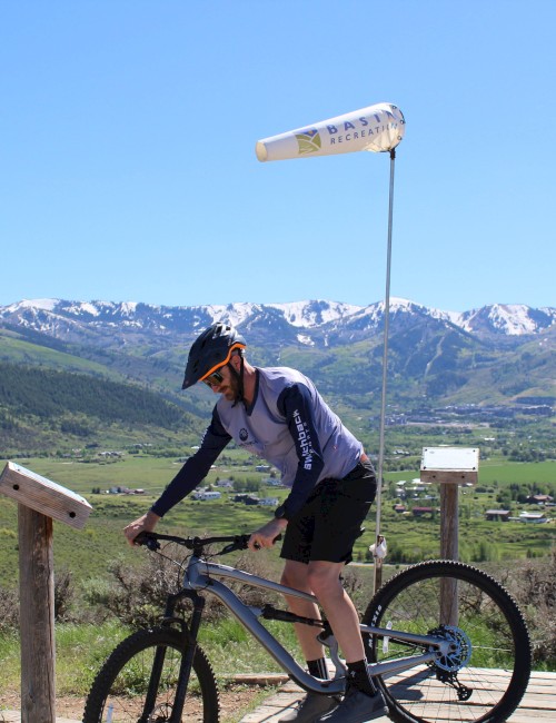 A person on a mountain bike, wearing a helmet and gloves, is stopped near a windsock on a trail with scenic mountains and a valley in the background.