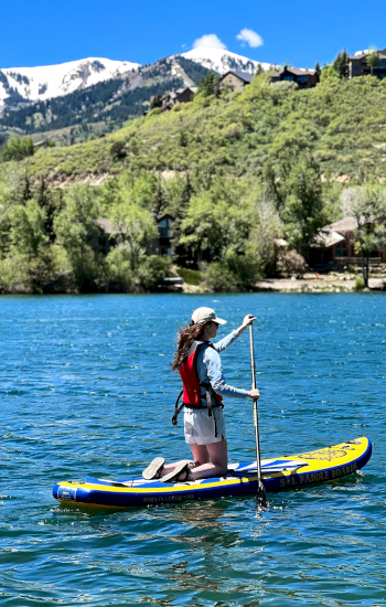 A person is paddleboarding on a serene body of water surrounded by green hills and snow-capped mountains, on a sunny day, wearing a life jacket.