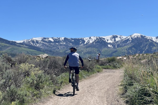 A person is riding a bike on a dirt trail through a scenic landscape with snow-capped mountains in the background under a clear blue sky.