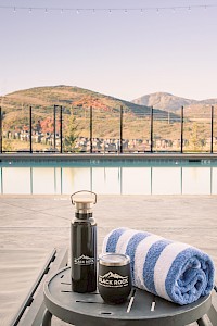 A poolside scene with a striped towel, water bottle, and container on a small table, overlooking mountains through a glass fence.
