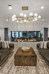 A modern living room features two brown sofas, a central wooden coffee table, a chandelier, a double-sided fireplace, and contemporary decor.