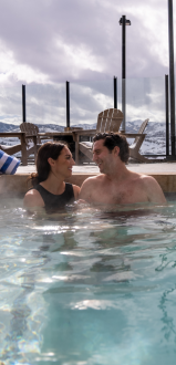 A couple is relaxing in a hot tub with mountains in the background. The scene is serene and outdoor furniture is visible.