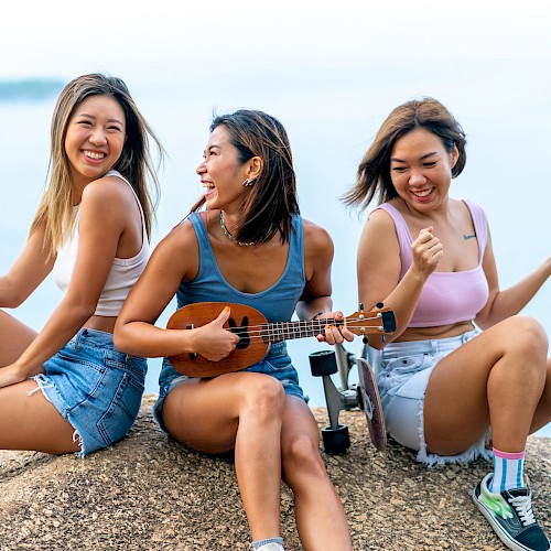 Three people sitting on a rock overlooking water, smiling and playing a ukulele.