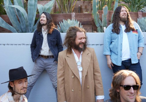 Five men are posing outdoors in front of plants; four are standing, and one is sitting. They're dressed in casual and semi-formal attire.