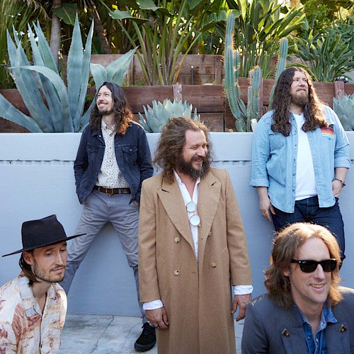 Five men are posing outdoors in front of plants; four are standing, and one is sitting. They're dressed in casual and semi-formal attire.