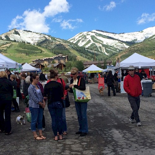 People are gathered at an outdoor market with white tents, surrounded by mountains partially covered in snow under a clear blue sky.