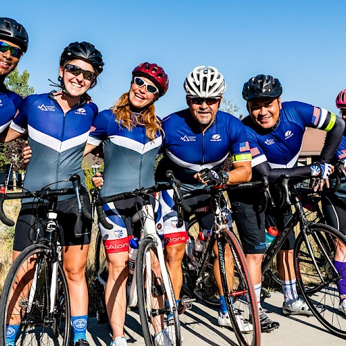 A group of six cyclists in blue jerseys pose with their bikes on a sunny day, smiling and ready for a ride.