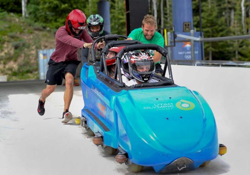 Two individuals are pushing a blue bobsled with two helmeted riders down a track at an outdoor facility, likely preparing for a ride.