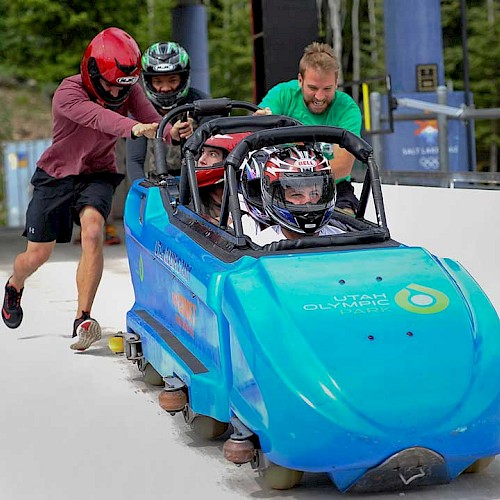 Two individuals are pushing a blue bobsled with two helmeted riders down a track at an outdoor facility, likely preparing for a ride.