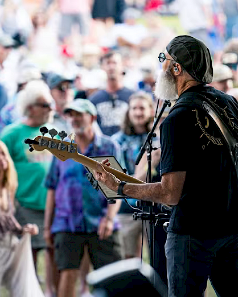A musician plays guitar on stage, facing an outdoor audience. The crowd appears engaged, with many people standing and watching the performance.