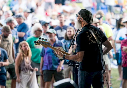 A musician plays guitar on stage, facing an outdoor audience. The crowd appears engaged, with many people standing and watching the performance.