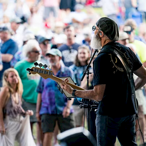 A musician plays guitar on stage, facing an outdoor audience. The crowd appears engaged, with many people standing and watching the performance.