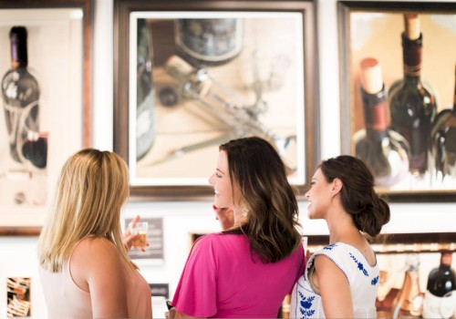 Three women are admiring wine-themed paintings in what appears to be an art gallery, smiling and conversing.