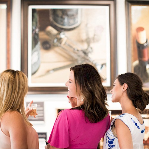 Three women are admiring wine-themed paintings in what appears to be an art gallery, smiling and conversing.