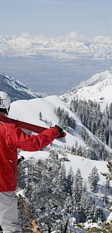 A skier in a red jacket holds skis, looking at a snowy mountain landscape with pine trees under a clear blue sky.