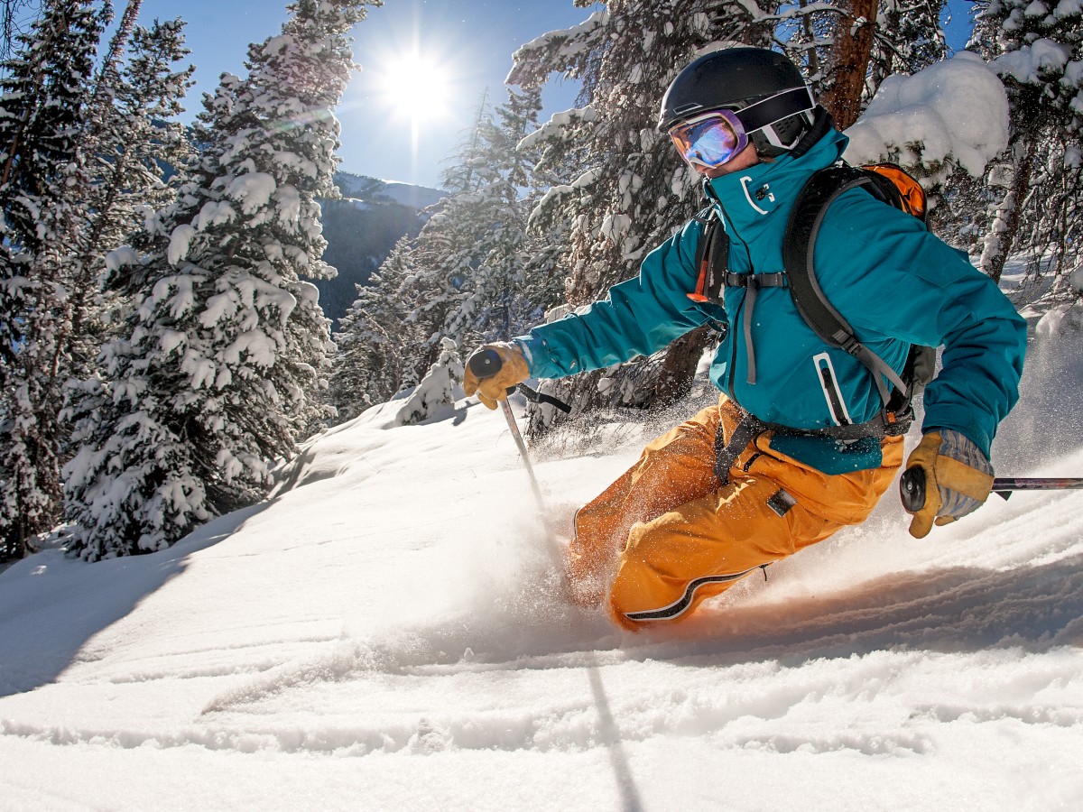 A skier wearing a blue jacket and orange pants is skiing down a snowy slope surrounded by trees with sunlight shining through.