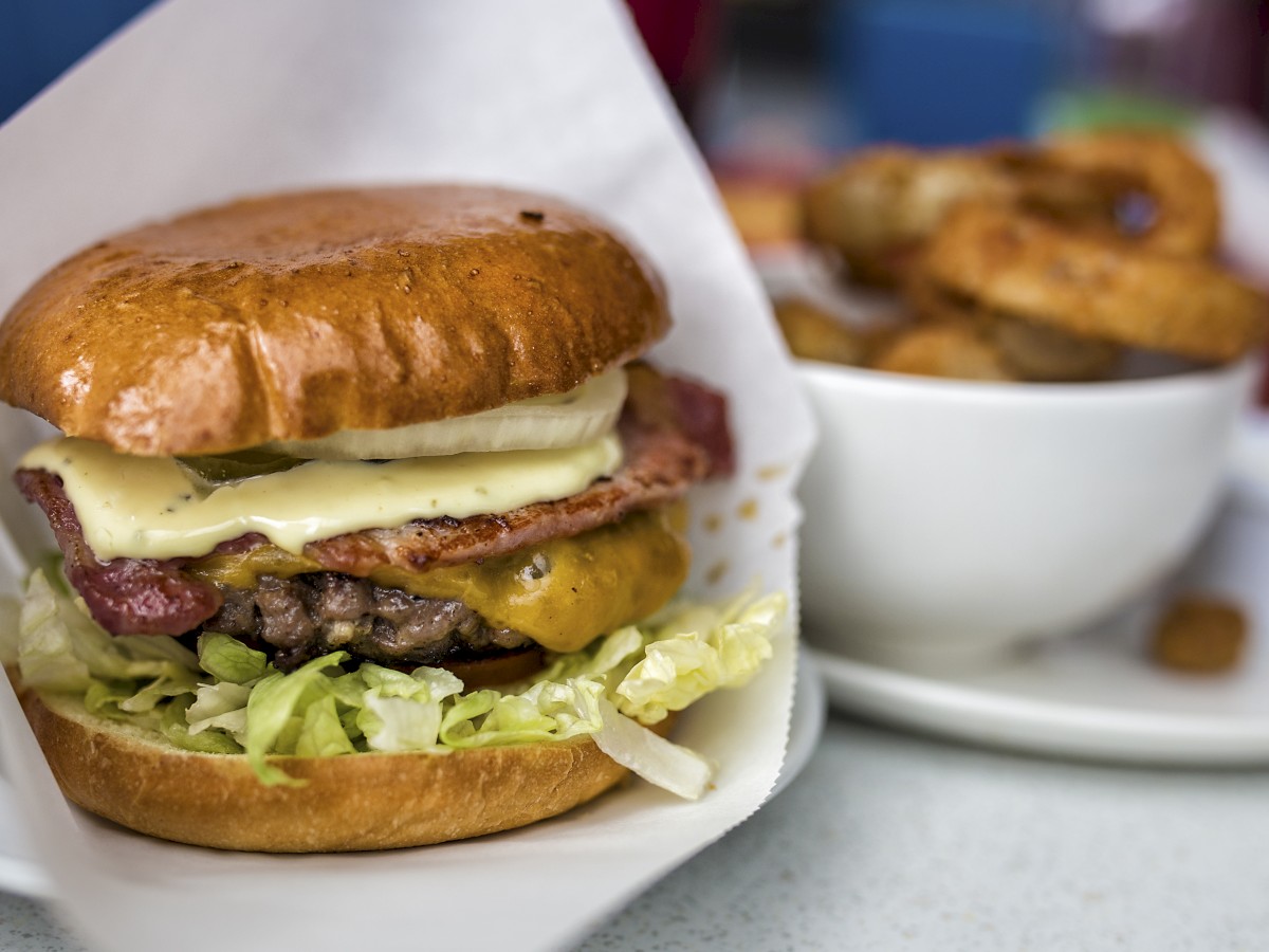 A cheeseburger with lettuce, bacon, and cheese in a bun sits next to a bowl of onion rings on a white table.