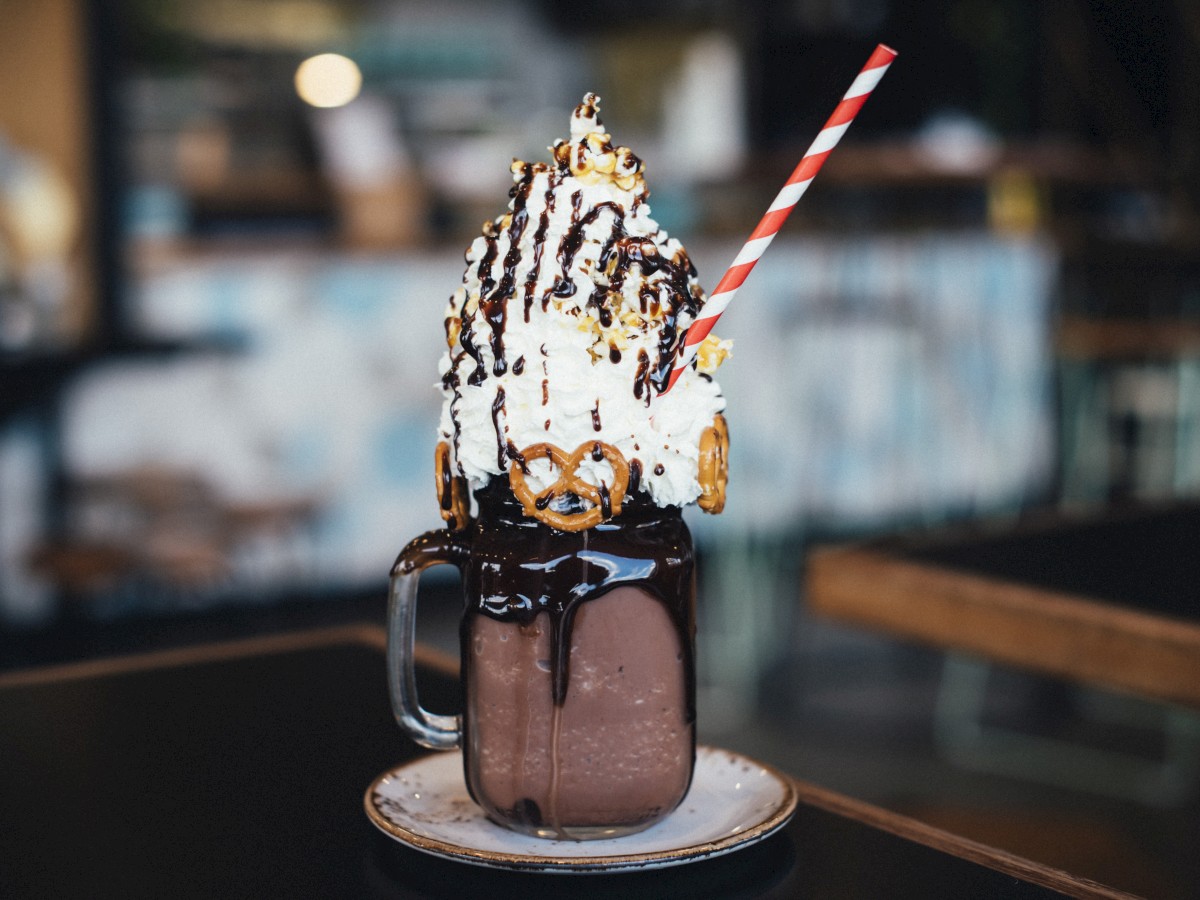 A decadent milkshake topped with whipped cream, pretzels, chocolate sauce, and a striped straw, served in a jar on a saucer.