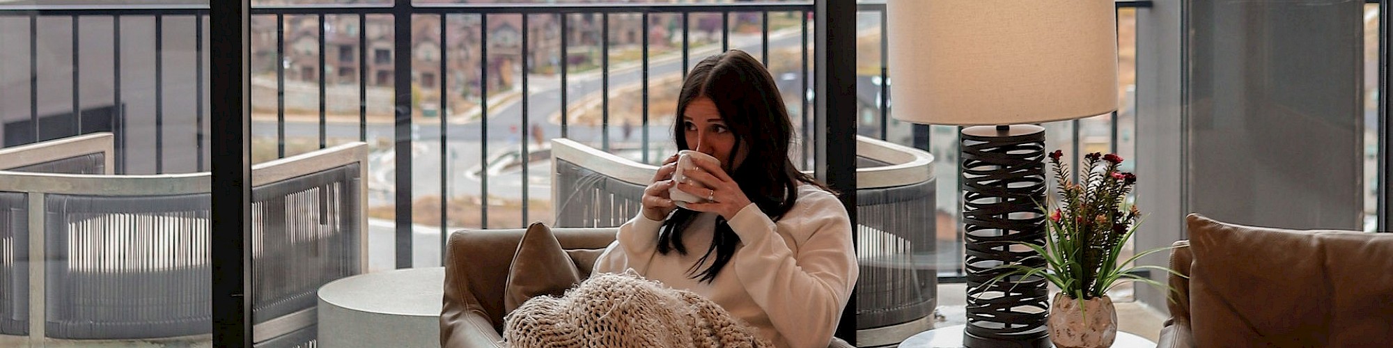 A person sits in a cozy room wrapped in a blanket, enjoying a drink with a scenic mountain view outside large windows.