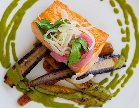 The image shows a beautifully plated dish featuring grilled salmon on top of roasted vegetables, garnished with greens and a green sauce drizzle.