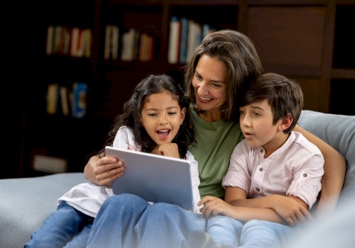 A woman and two children sit on a couch, happily looking at a tablet together, surrounded by a cozy living room with a bookshelf.