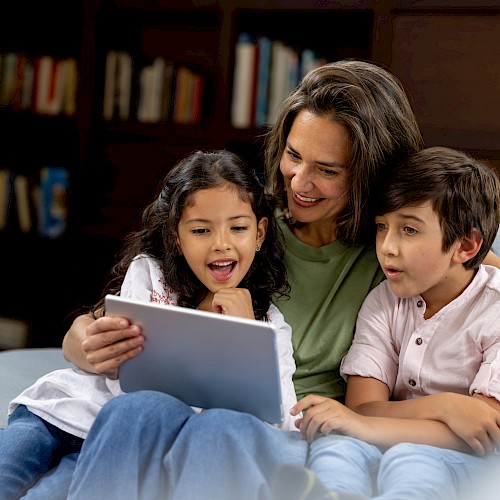 A woman and two children sit on a couch, happily looking at a tablet together, surrounded by a cozy living room with a bookshelf.