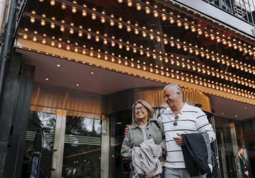 A couple is smiling and walking under a brightly lit marquee, possibly at a theater entrance.