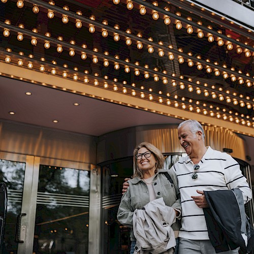 A couple is smiling and walking under a brightly lit marquee, possibly at a theater entrance.