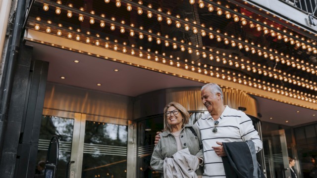 A couple is smiling and walking under a brightly lit marquee, possibly at a theater entrance.