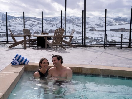 A couple sits in a rooftop hot tub with snowy mountains in the background, next to them is a rolled-up blue and white striped towel.