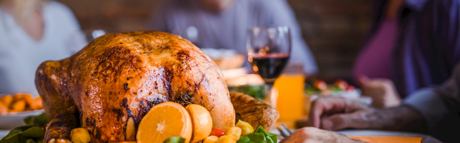 A roasted turkey garnished with oranges and vegetables, surrounded by people dining. A glass of red wine and a lit candle are visible.