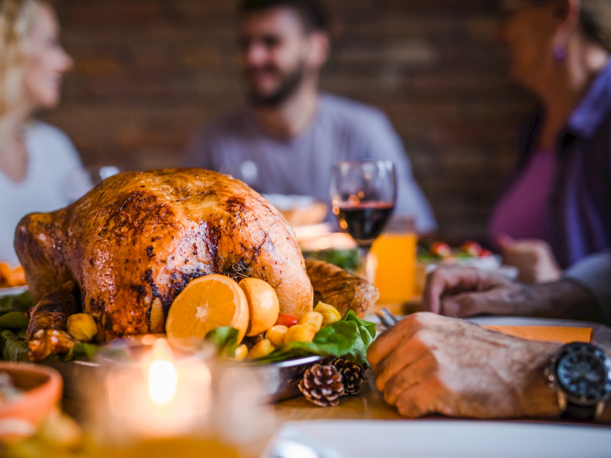 A roasted turkey garnished with oranges and vegetables on a dining table surrounded by people enjoying a meal together.