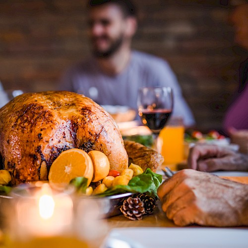 A roasted turkey garnished with oranges is on a table surrounded by people enjoying a meal together, with wine and candles present.