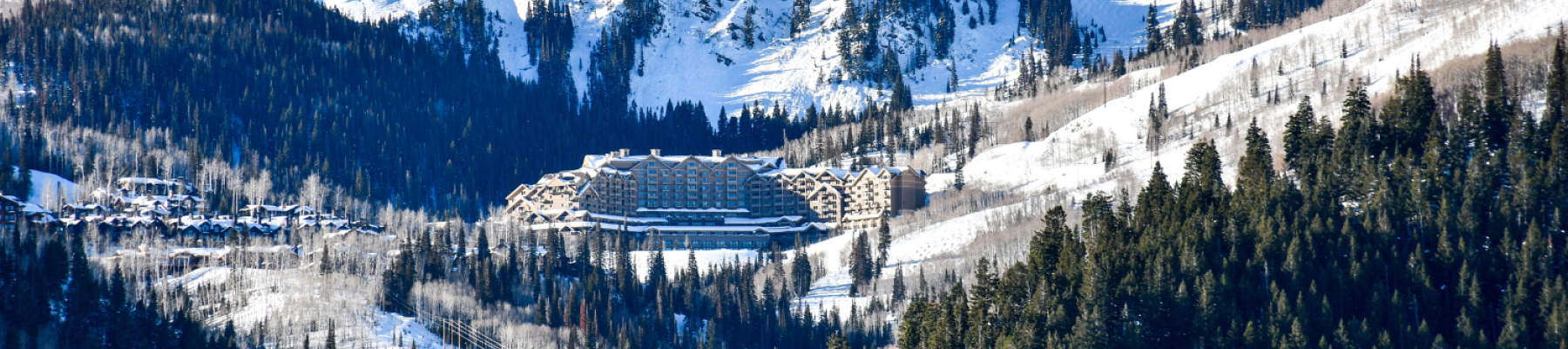 A snowy mountain landscape with a large resort nestled among trees, surrounded by snow-covered peaks and forested hillsides.