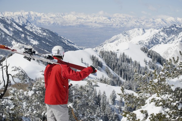 A skier in a red jacket holding skis over their shoulder looks at a scenic snowy mountain landscape.