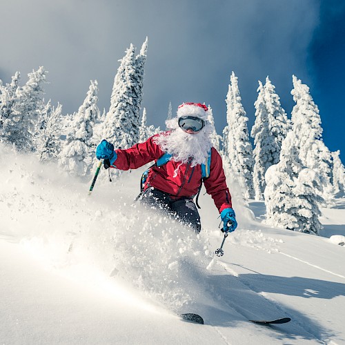 A skier dressed as Santa glides down a snowy slope surrounded by snow-covered trees and a clear blue sky.