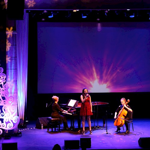 A musical performance on stage with a pianist, singer, and cellist, decorated with a white Christmas tree and a colorful backdrop.