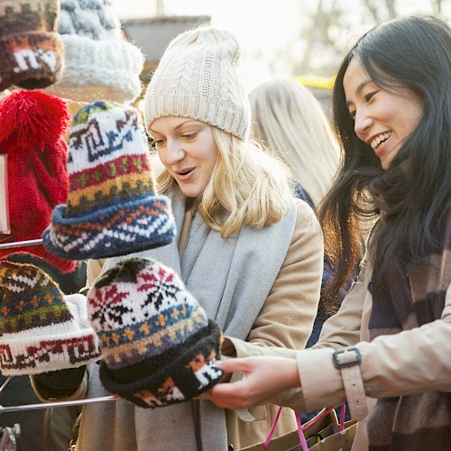 Two people are shopping for colorful and patterned winter hats at an outdoor market stall, both smiling as they browse.