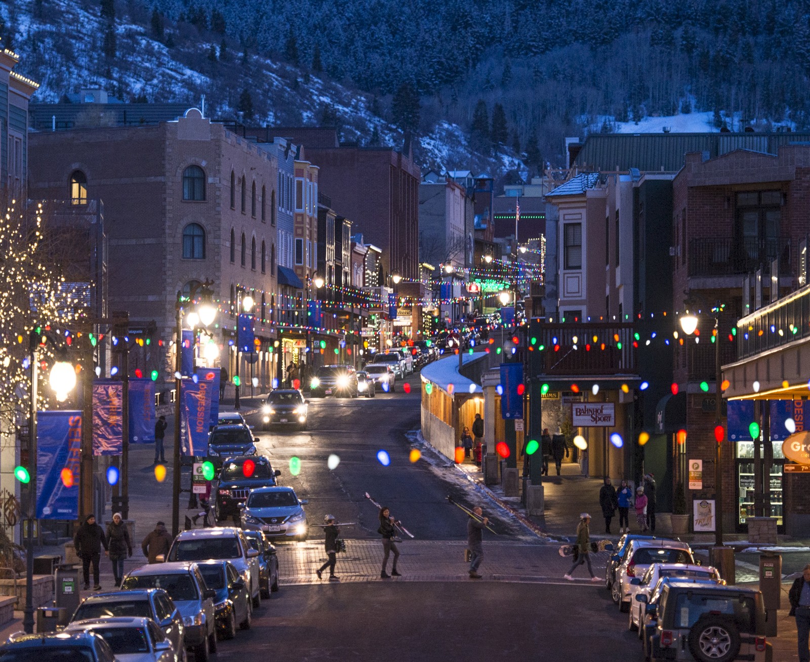 A festive street scene with colorful lights, parked cars, and people walking. The snow-covered backdrop adds to the wintery atmosphere.