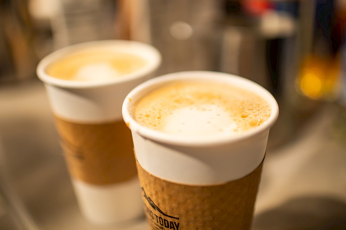 Two cups of coffee with frothy tops in paper cups, wrapped in brown sleeves, placed on a counter.