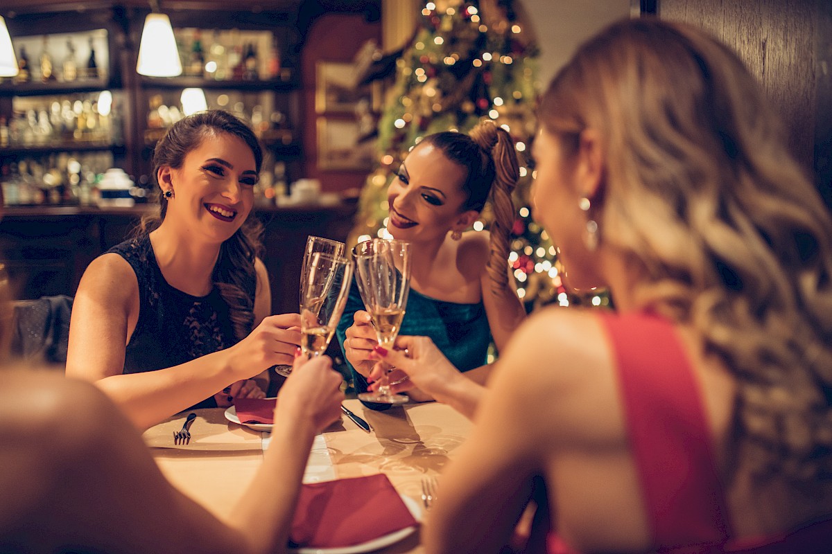 A group of women toasting with champagne glasses at a festive dinner table, with a Christmas tree in the background.