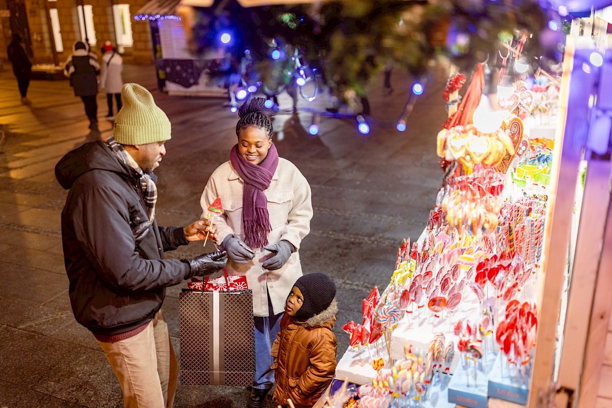 A family is at an outdoor market stall, admiring colorful candies and treats. It's a festive and cozy evening scene.