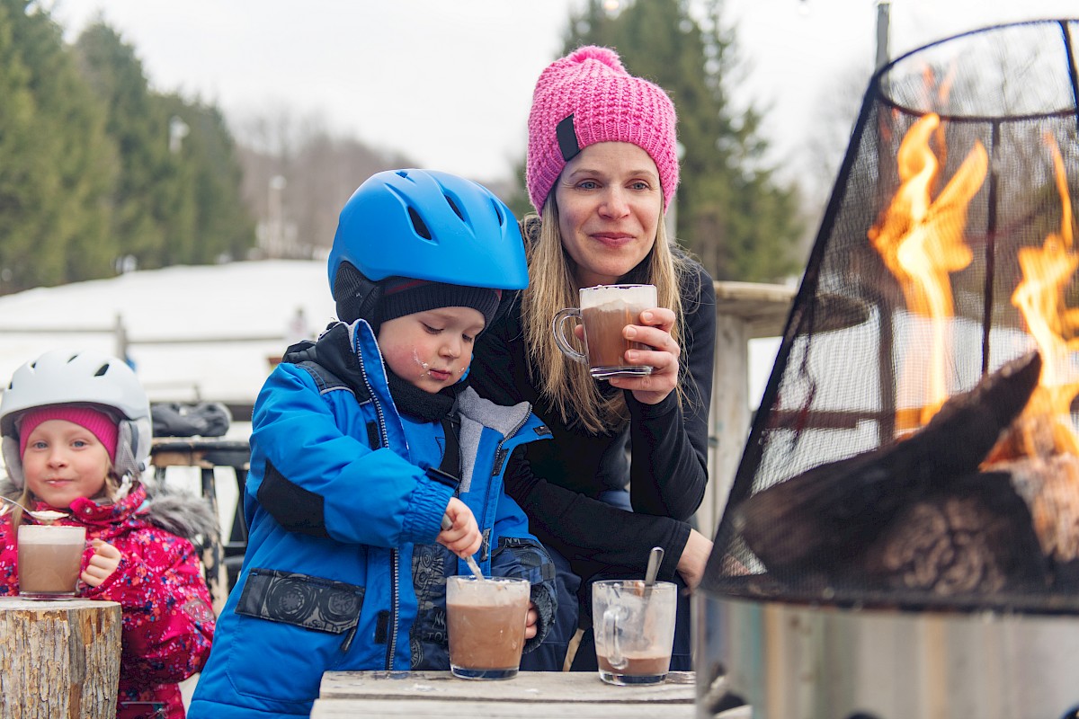 A person and two children enjoy hot drinks outdoors beside a fire. The children are wearing helmets and winter gear, sharing a cozy moment.