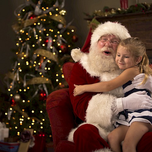 A child hugs a person dressed as Santa Claus, sitting in a festive chair near a decorated Christmas tree with lights and ornaments.