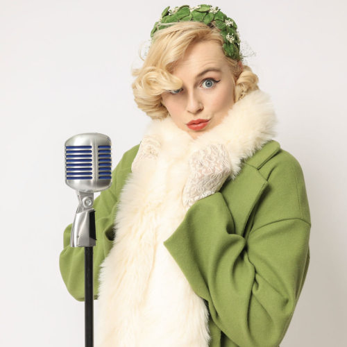 A person in vintage attire with a green coat and fur scarf stands by a retro microphone, giving a playful expression.