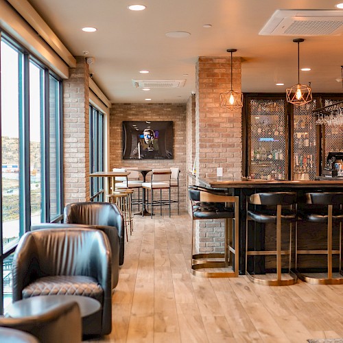 A modern bar area with wooden floors, high chairs, and comfortable seating. Large windows offer a scenic view outside.
