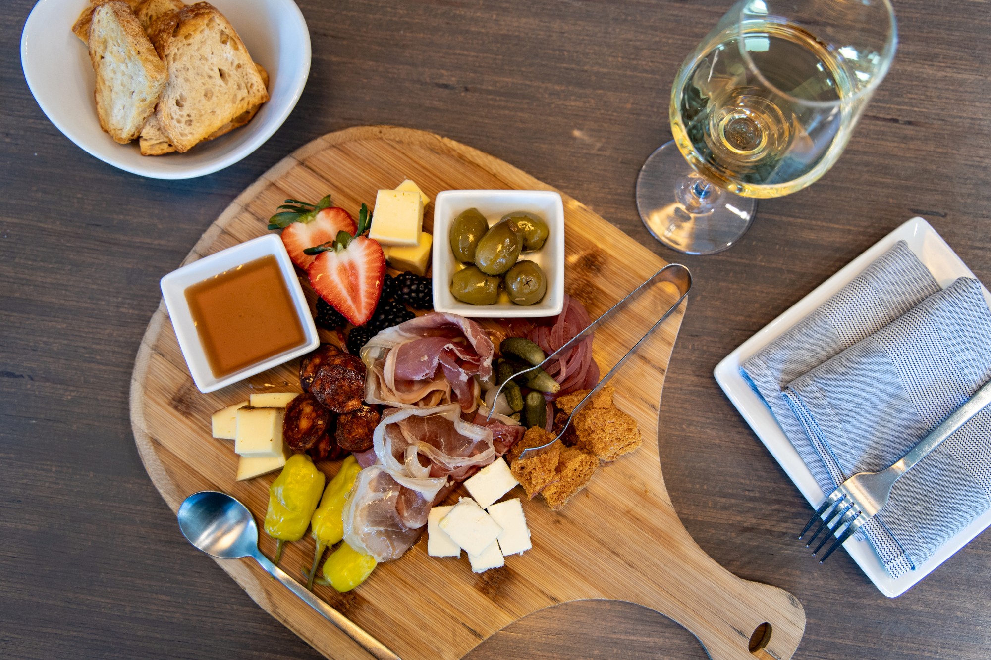 A charcuterie board with meats, cheeses, olives, and fruit; accompanied by bread and a glass of white wine.